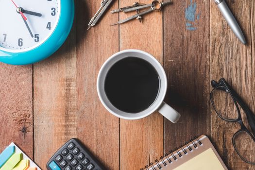 Top view of coffee cup with clock, glasses and office supplies on wooden table background.