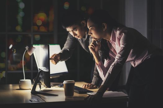 Asian businesswoman and businessman working hard late together with technology computer in office, customer service and call center, team work with colleagues for success achievement project concept