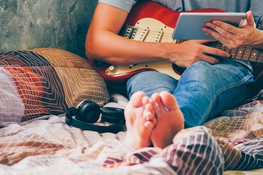 Close up of young man using tablet and playing electric guitar on his bed, Films grain filter.