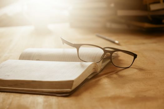 Close up of open notebook with eyeglasses on wooden desk, Soft focus, Flare sun light,Vintage tone.
