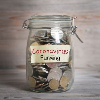 Coins in glass money jar with Coronavirus Funding label, financial concept. Vintage wooden background with dramatic light.