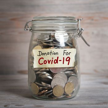 Coins in glass money jar with donation for covid19 label, financial concept. Vintage wooden background with dramatic light.