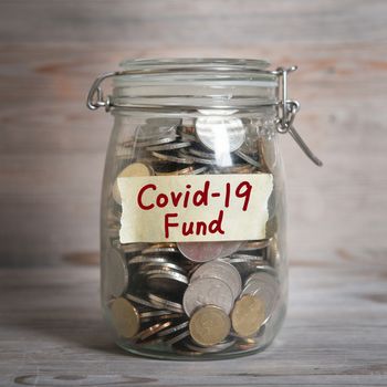 Coins in glass money jar with covid19 fund label. Vintage wooden background with dramatic light.