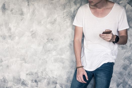 Young man wearing white t-shirt and blue jeans, holding smart phone and standing on gray grunge background. Free space for text