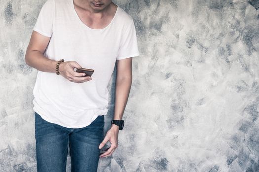 Young man wearing white t-shirt and blue jeans, holding smart phone and standing on gray grunge background. Free space for text
