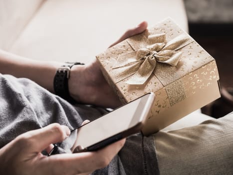 Close up of casual man's hands using smart phone and holding gift box.