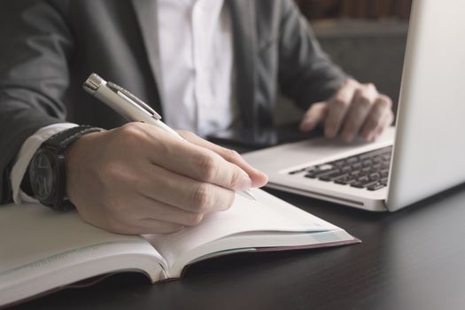 Close up of businessman writing note and using a laptop on the office desk.