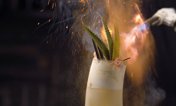 Close up bartenders hands sprinkling with burning cinnamon pina colada cocktail. Glass on the background of the fire. Bar drinks series.