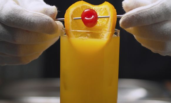 Making a Tequila Sunrise Cocktail. Close up bartenders hands garnish long drink with orange and cherry on skewer.