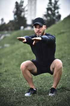 Young caucasian man performing squats before jogging on footpath in park.