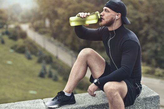 Muscular bearded athlete drink a water after good workout session on city park.