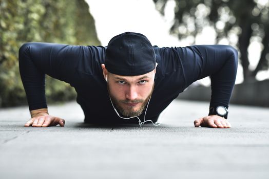 Young bearded sportsman makes workout in a park, doing a series of push ups outdoor.