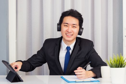 Work from home, Asian young businessman wear suit video conference call or facetime he smiling sitting on desk touch smart digital tablet computer and write paper notebook information at home office