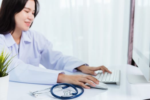 Asian beautiful female doctor smiling wearing doctor's uniform with stethoscope typing on keyboard desktop computer looking to monitor on the desk at hospital office, Medical healthcare concept