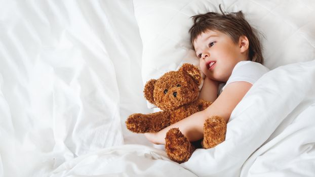 Toddler lies in bed with cute teddy bear. Little boy under white blanket with fluffy toy. Plush guard watches out child's sleep. Background with copy space.