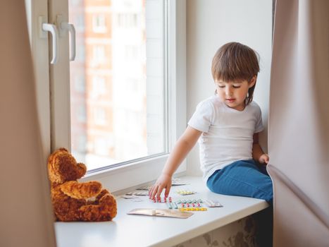 Toddler sits on windowsill and plays with scattering pills without parent's control. Dangerous situation with little boy. Medicines are freely available to child.