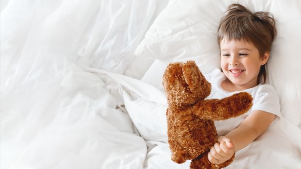 Toddler lies in bed with cute teddy bear. Little boy under white blanket with fluffy toy. Plush guard watches out child's sleep. Morning bedtime at cozy home.