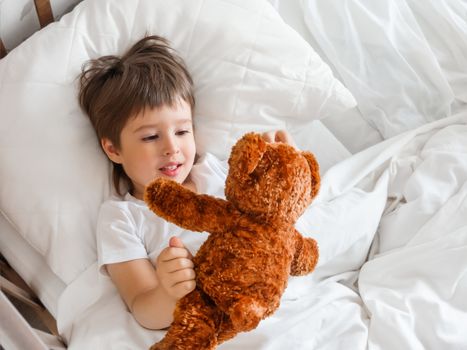 Toddler lies in bed with cute teddy bear. Little boy under white blanket with fluffy toy. Plush guard watches out child's sleep. Morning bedtime at cozy home.