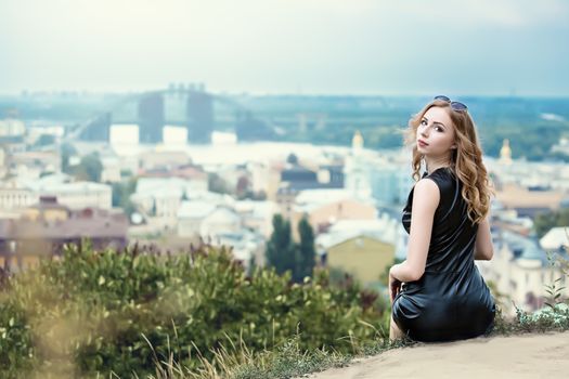 The beautiful lonely girl in black dress and sunglassess sitting on the ground and looking back