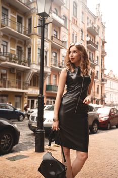 Stylish, luxurious girl in a chic black dress on the street, female with fashion makeup near the building, black dress on tall woman with long legs walks in the city.