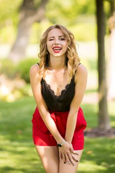 Attractive funny young woman showing tongue and winking eye, beautiful female with curly hair looking at camera, flirting, pretty girl wearing black T-shirt posing in park