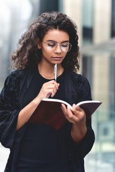 Charming Afro American beautiful happy female, making notes in notepad while walking outdoors on a city street. Education and business concept with space for text.