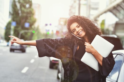 Young happy black girl try to take a taxi on the street holding a laptop