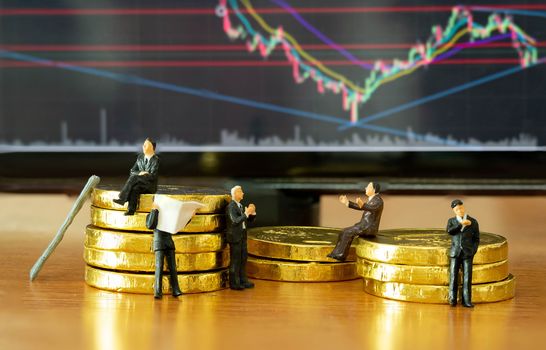 The miniature figure a group of business men sitting on top of pile stack of golden coins and some standing reading newspaper and discuss to other  with a Stock Market Techinical Graph Candlestick Background business success concept