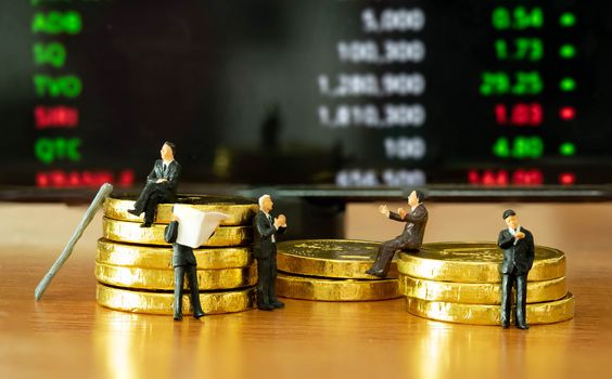 The miniature figure a group of business men sitting on top of pile stack of golden coins and some standing reading newspaper and discuss to other  with a Stock Market Techinical Graph Ticker Background business success concept