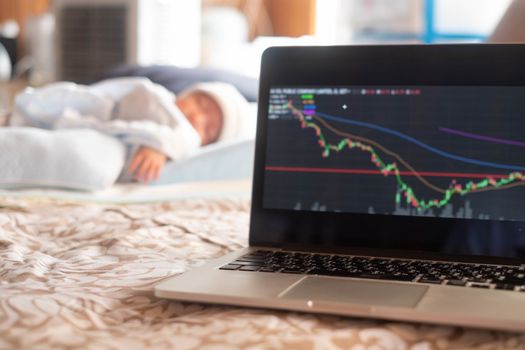 Soft focus on laptop stock display screen .The parents father Stock trader trading stock on his laptop while his newborn infant baby sleep on the bed