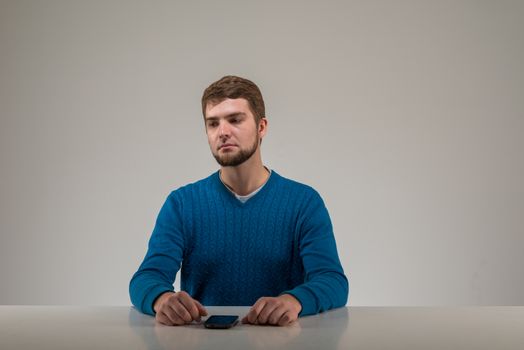 Sad bearded young man wearing blue sweater sits at the table with his phone