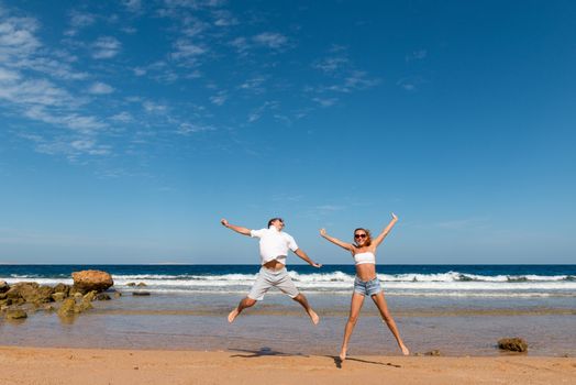 Happy couple jumping on the beach excited with wave behind