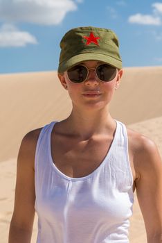 Beautiful young woman wearing sunglasses and military cap with red star in desert sand dunes