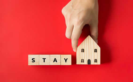 Hand hold on to the house model and text the word "Stay" on the wooden dice. In the concept, stay at home to stop the spread. During the corona virus outbreak or COVID-19 on red background