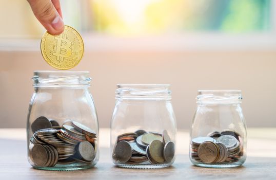 Money savings concepts hand holding bitcoin to put with coins in glass bottles to spend on expenses such as savings, tourism, investment, emergency, retirement on wooden table with blur background 