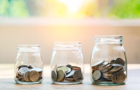 Money savings concepts. Put coins in glass bottles to spend on expenses such as savings, tourism, investment, emergency, retirement on wooden table with blur bokeh background with copy space