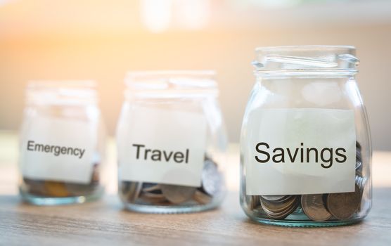 Money savings concepts. Put coins in glass bottles with paper label and text to spend on expenses such as savings, tourism, investment, emergency, retirement on wooden table with blur background