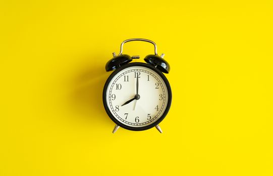 black alarm clock from 7 a.m. to 8 a.m. with bright yellow  background and shadows from the changing light of the sun's movements in morning and working hours concept front shot