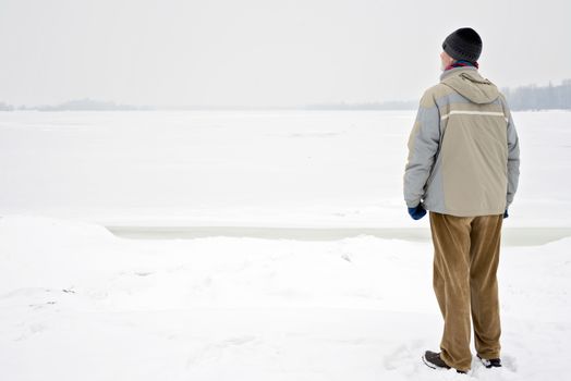 A man with a wool cap, a waterproof jacket and a wool cap is looking at the Dnieper river during a cold and sad gray winter morning under the snow