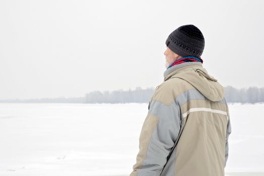 A man with a wool cap, a waterproof jacket and a wool cap is looking at the Dnieper river during a cold and sad gray winter morning under the snow