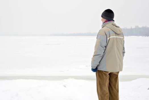 A man with a wool cap, a waterproof jacket and a wool cap is looking at the Dnieper river during a cold and sad gray winter morning under the snow