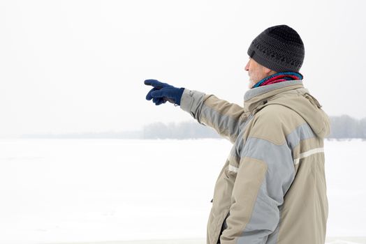 A man with a wool cap, a waterproof jacket and a wool cap is indicating something with the finger close to the Dnieper river during a cold and sad gray winter morning under the snow