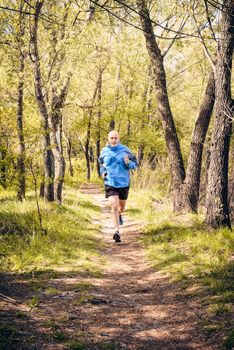 A senior man dressed in black and blue is running in the forest, during a warm spring day