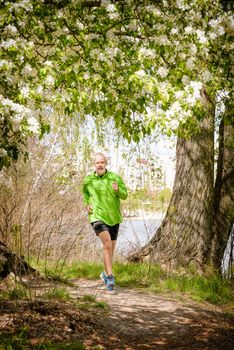 A senior man dressed in black and green is running in the forest, close to the lake, under an apple blossom, during a warm spring day