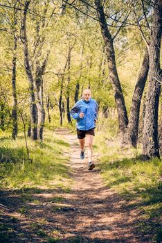 A senior man dressed in black and blue is running in the forest, during a warm spring day