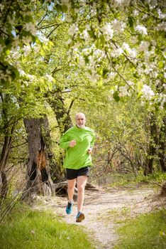 A senior man dressed in black and green is running in the forest during a warm spring day