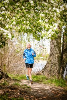 A senior man dressed in black and blue is running in the forest, close to the lake, under an apple blossom, during a warm spring day