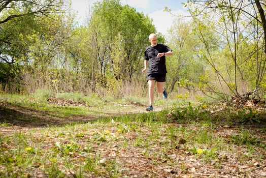 A senior man dressed in black is running in the forest, and looks at the time on his stopwatch, during a warm spring day