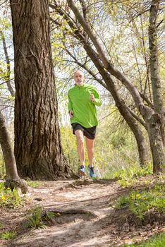 A senior man dressed in black and green is running in the forest, during a warm spring day