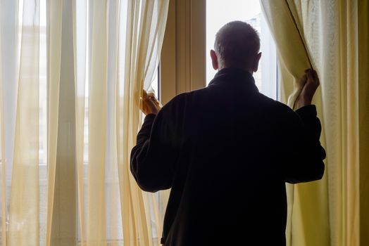 A man in black moves the curtain aside to look out trough the window